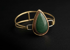 Side profile view of a handcrafted mixed-metal bangle bracelet showcasing a teardrop green aventurine gemstone in a copper crown bezel, open brass wire band, and silver clasp loops resting on a dark surface.