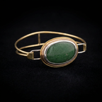 Side profile view of a handcrafted mixed-metal oval bangle bracelet showcasing a smooth green aventurine gemstone in a multi-tonal bezel, double brass wire band, and silver clasp loop on a dark background.