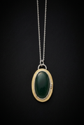 Handcrafted oval green aventurine pendant featuring a smooth cabochon gemstone set in a patinated brass bezel frame with silver accent beads and top bail loop resting on a dark moody surface.