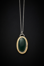 Handcrafted oval green aventurine pendant featuring a smooth cabochon gemstone set in a patinated brass bezel frame with silver accent beads and top bail loop resting on a dark moody surface.