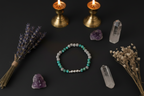 A beaded bracelet with turquoise, gray, and white beads is arranged on a dark surface with crystals, dried lavender, and two lit brass candles as styled props.