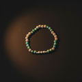 A beaded bracelet made of green and tan textured round beads with a short section of small silver metallic beads, photographed on a dark brown background with a spotlight effect.