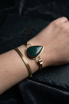 A gold-toned cuff bracelet with a large teardrop-shaped dark green stone, worn on a person's wrist alongside a thinner gold bangle.