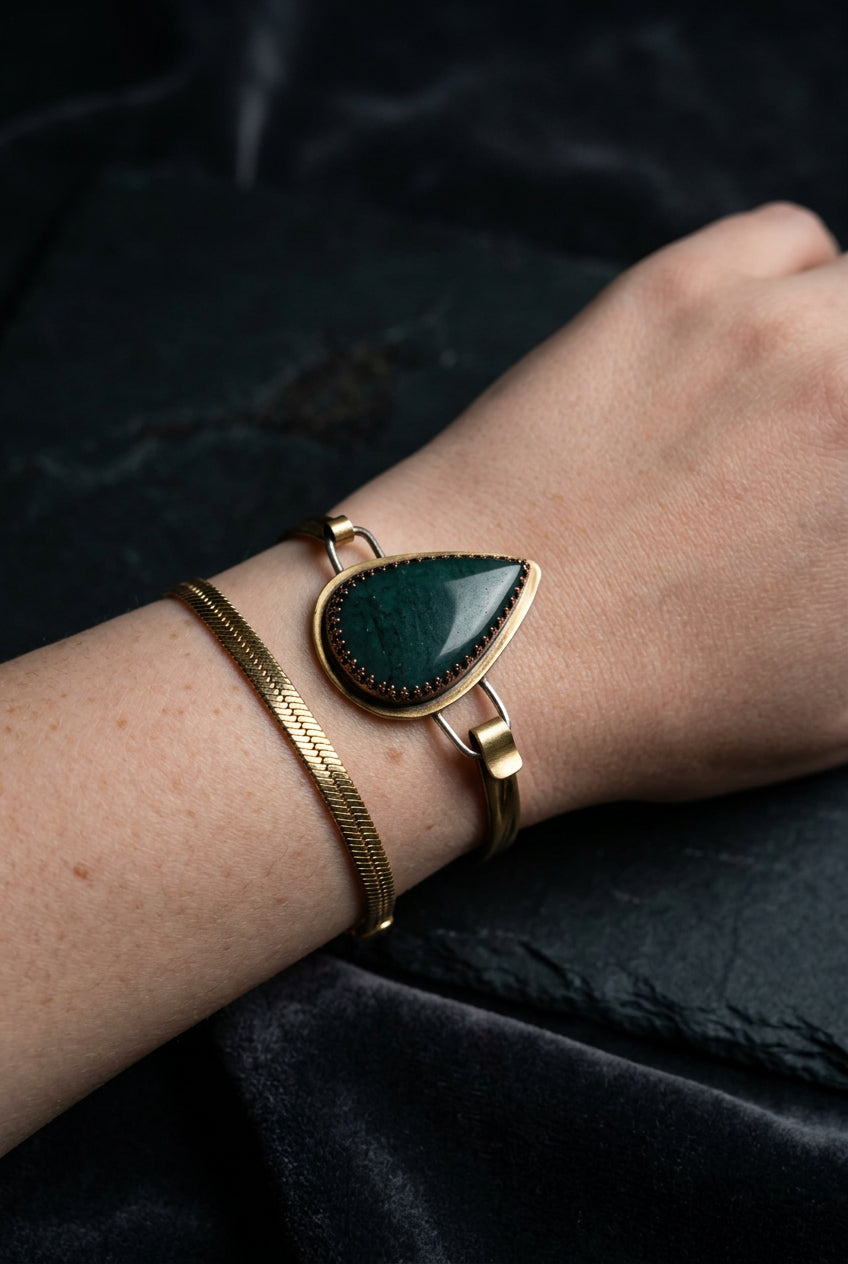 A gold-toned cuff bracelet with a large teardrop-shaped dark green stone, worn on a person's wrist alongside a thinner gold bangle.