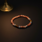 A beaded stretch bracelet made of peach and pink round beads, accented with textured white, brown, and silver beads, displayed on a dark surface with a warm brass object in the background.