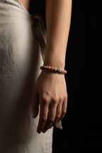 A close-up of a person's wrist wearing a beaded bracelet with orange, brown, and gray beads, paired with a light beige linen dress against a dark background.