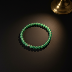 A green beaded bracelet with a small silver accent bead, displayed on a dark surface with a warm spotlight and a brass object in the top right corner.