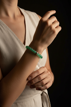 A close-up of a person wearing a green beaded bracelet on their wrist, dressed in a light beige sleeveless top against a dark background.