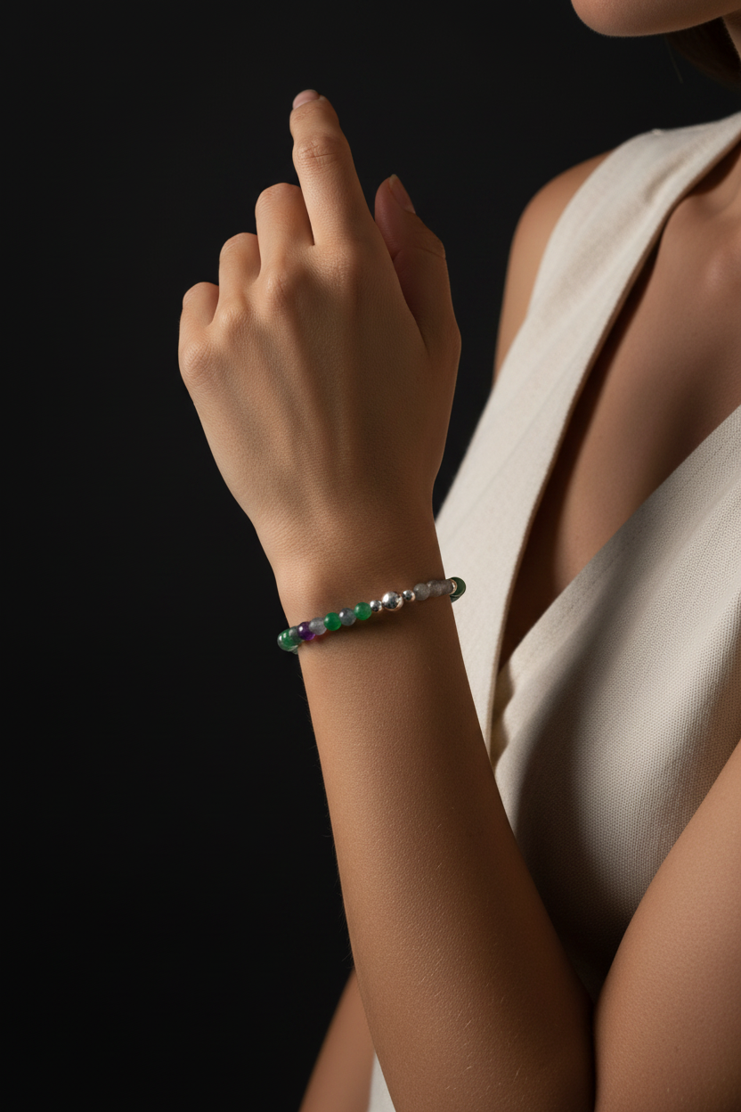 A close-up lifestyle shot of a woman's hand wearing a beaded bracelet with green, purple, and silver beads, set against a dark background and her cream-colored sleeveless top.
