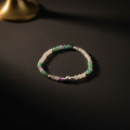 A beaded stretch bracelet featuring translucent white beads, green beads, purple beads, and metallic silver beads, displayed on a dark textured surface.