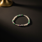 A beaded stretch bracelet featuring translucent white beads, green beads, purple beads, and metallic silver beads, displayed on a dark textured surface.