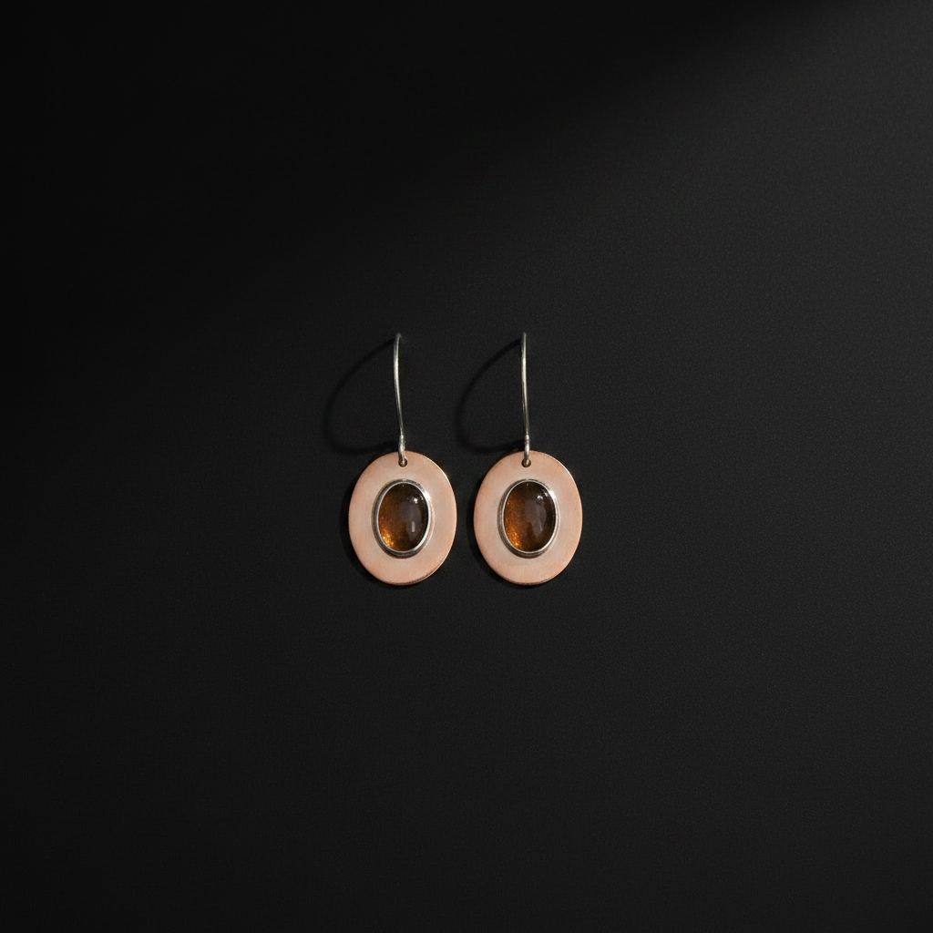 A pair of minimalist dangle earrings featuring circular peach-pink metal discs with a central oval amber-brown stone, suspended from thin silver-toned hooks against a dark black background.