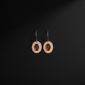 A pair of minimalist dangle earrings featuring circular peach-pink metal discs with a central oval amber-brown stone, suspended from thin silver-toned hooks against a dark black background.