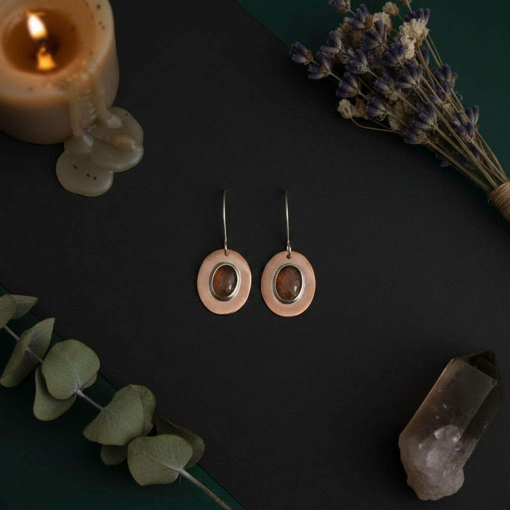 A pair of handmade dangle earrings with circular rose gold-toned outer rings, smooth brown stone centers, and silver-toned hooks, displayed on a dark background with candles, dried flowers, eucalyptus leaves, and a crystal.