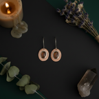 A pair of handmade dangle earrings with circular rose gold-toned outer rings, smooth brown stone centers, and silver-toned hooks, displayed on a dark background with candles, dried flowers, eucalyptus leaves, and a crystal.