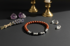 A beaded stretch bracelet with terracotta orange, white, and black beads, styled on a dark gray surface with lavender, crystals, and brass candlesticks in the background.