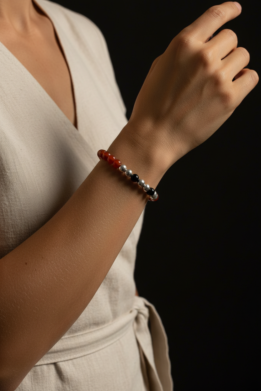 A close-up of a person's wrist wearing a beaded stretch bracelet with red, silver, and dark blue beads, styled with a cream-colored wrap dress against a solid black background.