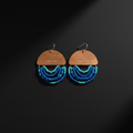 A pair of dangling earrings with wooden semi-circular tops engraved with 'ENOUGH', hanging silver hooks, and multiple curved rows of blue and turquoise beads below, displayed on a dark background.