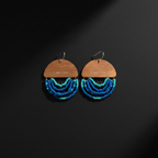 A pair of dangling earrings with wooden semi-circular tops engraved with 'ENOUGH', hanging silver hooks, and multiple curved rows of blue and turquoise beads below, displayed on a dark background.
