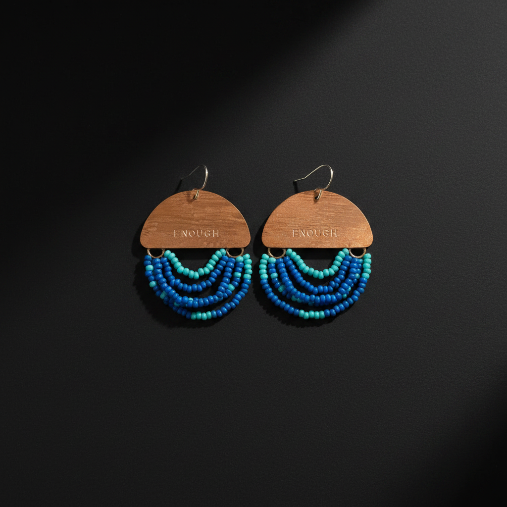 A pair of dangling earrings with wooden semi-circular tops engraved with 'ENOUGH', hanging silver hooks, and multiple curved rows of blue and turquoise beads below, displayed on a dark background.