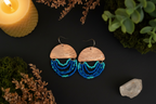 A pair of handmade bohemian earrings featuring a light wood semicircle top and cascading blue and teal beaded fringe, displayed on a dark background with decorative props including a lit candle, moss, dried flowers, and a crystal.