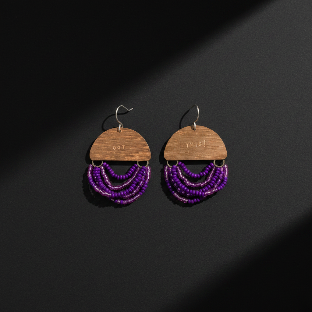 A pair of handmade dangle earrings featuring a semi-circular wooden top with engraved text, hanging from silver fishhook wires, and adorned with multiple layers of purple beaded fringe, displayed on a dark background with dramatic diagonal shadows.