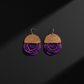 A pair of handmade dangle earrings featuring a semi-circular wooden top with engraved text, hanging from silver fishhook wires, and adorned with multiple layers of purple beaded fringe, displayed on a dark background with dramatic diagonal shadows.