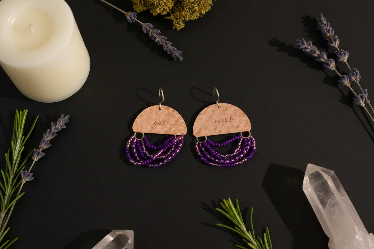 A pair of handmade dangle earrings with light wood semicircle tops, silver hooks, and multiple strands of purple beads, displayed on a dark background with candles, lavender, rosemary, and crystal points.