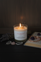 A lit white candle in a clear glass jar sits on a dark tabletop with dried lavender, crystals, and an open book nearby. The candle label reads 'Gratitude' and 'Scented Candles'.