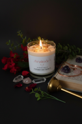 A lit white candle in a clear glass jar with a white label reading 'Gratitude' and 'Wiccan Candle,' styled on a dark surface with red flowers, green leaves, crystals, an open book, and a brass tool.