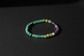 A beaded stretch bracelet made of alternating translucent green, pale yellow, white, and purple beads, displayed on a dark black background.