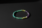 A beaded stretch bracelet made of alternating translucent green, pale yellow, white, and purple beads, displayed on a dark black background.