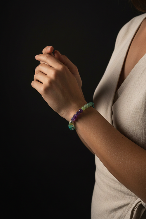 A close-up of a person's hand and wrist wearing a beaded stretch bracelet with green, purple, and blue beads, paired with a beige sleeveless top against a solid black background.