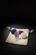 Four gemstones and a white tumbled sphere displayed on a small cream drawstring pouch with a printed logo, set against a dark black background.