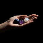 Four polished tumbled crystals in dark blue, brown, purple, and white are held in a person's open palm against a solid black background.