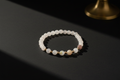 A beaded stretch bracelet featuring white and pale pinkish-beige round beads separated by small gold spacer beads, displayed on a dark surface with dramatic lighting.