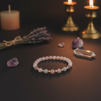 A beaded stretch bracelet with pale pink, mint green, and white beads separated by small gold spacer beads, displayed on a dark surface with crystals, lavender, and lit candles in the background.