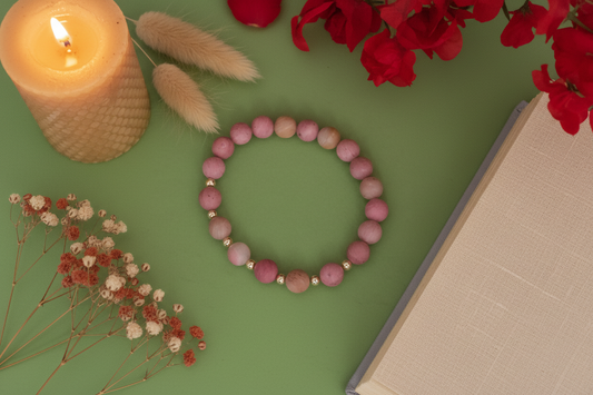Pink beaded bracelet on a green surface with a lit candle, dried flowers, and red roses.