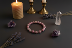 A pink beaded bracelet with small gold spacer beads is arranged on a dark gray surface, surrounded by decorative props including a lit cream candle in a brass holder, lavender sprigs, a clear crystal point, and purple mineral stones.