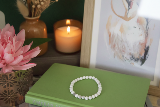 White beaded bracelet on a green book with a pink flower, candle, and framed picture in the background.