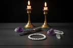 A white beaded bracelet arranged on a dark wooden surface, surrounded by crystals, a bundle of dried lavender, and two lit gold candlesticks against a black background.