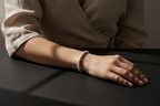 A close-up of a person's hand resting on a dark surface, wearing a delicate white beaded bracelet. The person is wearing a beige linen wrap top, and the scene is lit by dramatic, directional sunlight casting soft shadows.