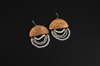 A pair of handmade dangle earrings featuring engraved wooden semicircles with the word 'Light' on them, hanging from gold-toned fishhook wires. Below the wood pieces are multiple layers of white and silver beaded fringe, set against a solid black background.