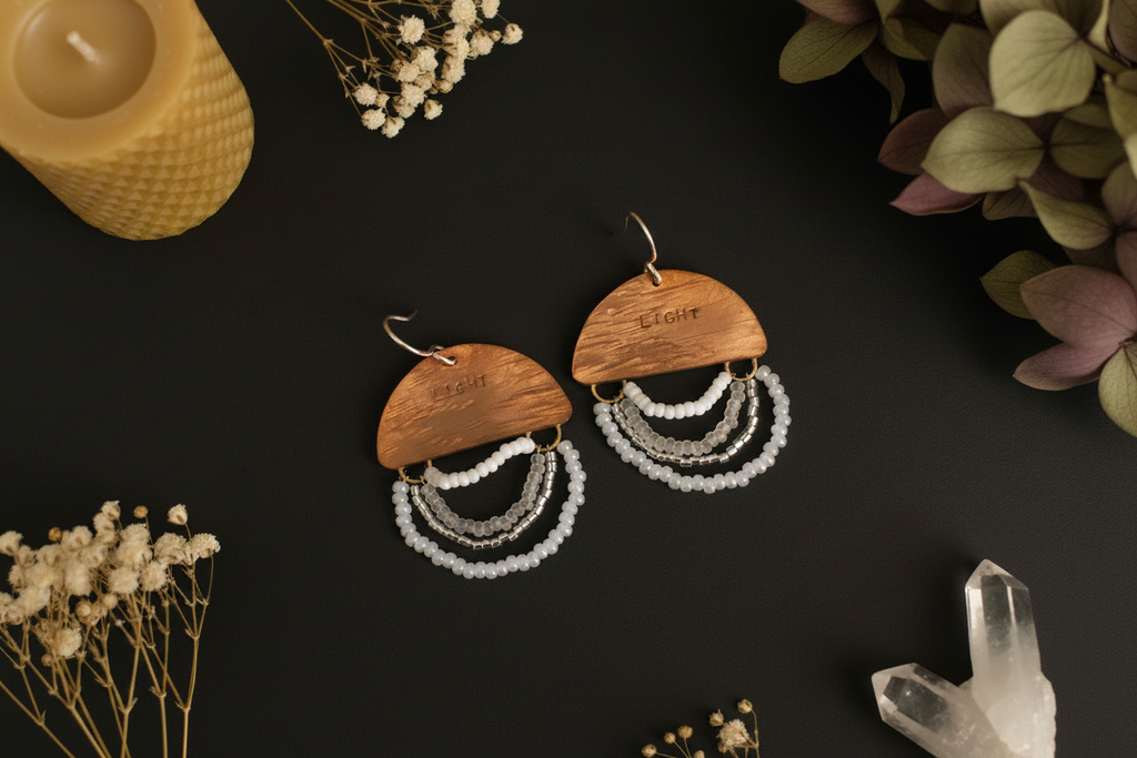 A pair of bohemian-style wooden dangle earrings with white and silver beaded fringe, displayed on a dark background with dried flowers and a candle.