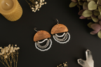 A pair of bohemian-style wooden dangle earrings with white and silver beaded fringe, displayed on a dark background with dried flowers and a candle.
