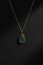A silver chain necklace with a teardrop-shaped pendant featuring a blue iridescent stone and a gold-toned bezel setting, displayed against a dark background.