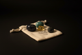 Four polished gemstone crystals in black, teal, and yellow, displayed on a small cream-colored drawstring pouch with a printed logo, set against a solid black background.