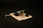Four polished gemstone crystals in black, teal, and yellow, displayed on a small cream-colored drawstring pouch with a printed logo, set against a solid black background.