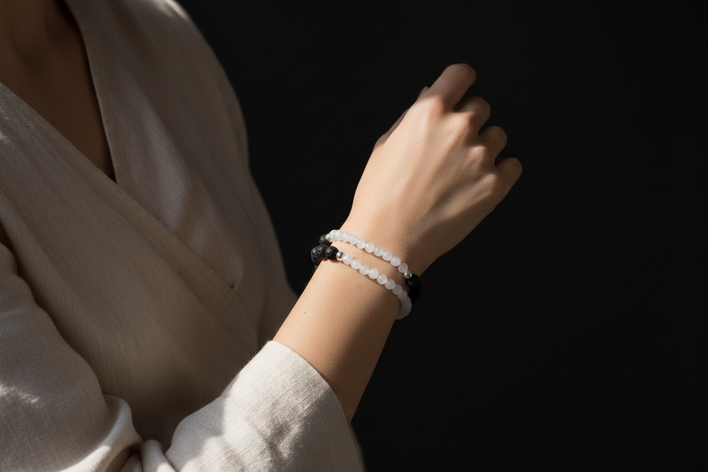 A close-up of a person's hand and wrist wearing two beaded bracelets against a dark background. One bracelet is white with small black beads, and the other is white with larger black beads and a black central connector.