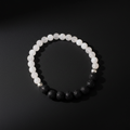 A beaded stretch bracelet arranged in a circle on a dark background, featuring translucent white round beads on one side and matte black porous round beads on the other, with a small metallic silver bead near the top right.